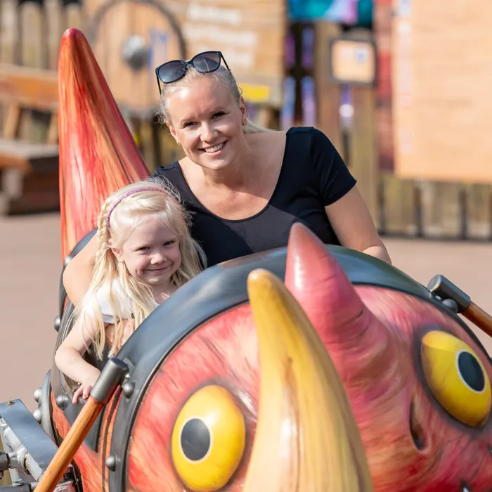 Eine Frau und ein fröhliches Mädchen sitzen in der Gondel von Grobians Wolkenspringer im Heide Park Resort. Die Gondeln sind bunte Drachen.