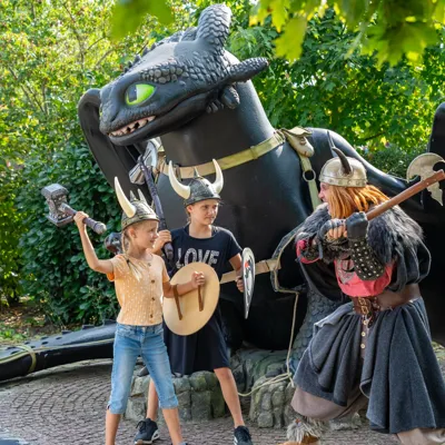 Ein Wikinger steht mit zwei Mädchen mit Wikinger-Utensilien vor einer großen Ohnezahn-Drachen-Figur