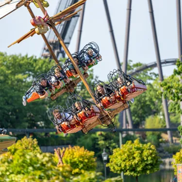 Besucher schweben mit der Attraktion "Hicks Himmelstürmer" im Heide Park Resort liegend durch die Lüfte. Im Hintergrund sind Bäume zu sehen und die Attraktion KRAKE.