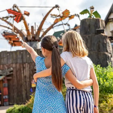 Zwei Mädchen haben die Arme umeinander gelegt. Sie zeigen und schauen in die Ferne. Dort ist "Hicks Himmelsstürmer" zu sehen. Eine Attraktion im Heide Park Resort. Besucher schweben liegend durch die Luft